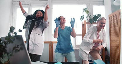 Zoom out three fun happy diverse doctor women dance together celebrating victory over virus pandemic at clinic lab.