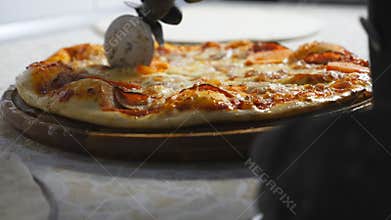 Male hand of chef in glove cutting tasty pizza into slices with a round cutter knife on a wooden board. Arm of cook cuts