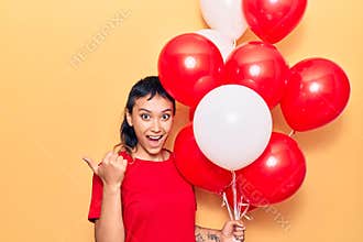 Young woman holding balloons pointing thumb up to the side smiling happy with open mouth