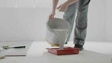 Woman builder pours white paint into red tray in apartment