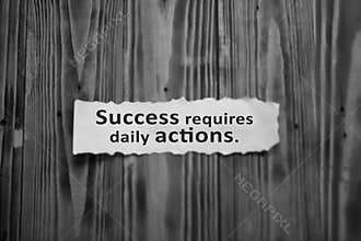 Success requires daily actions. Inspirational words on white torn paper note on wooden wall background in black and white.