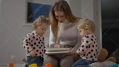 Absorbed millennial mother surfing Internet on tablet as twin daughters standing around. Portrait of slim beautiful