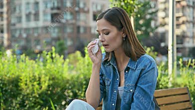 Young woman sneezing into tissues, wiping nose, sitting on bench, outdoor
