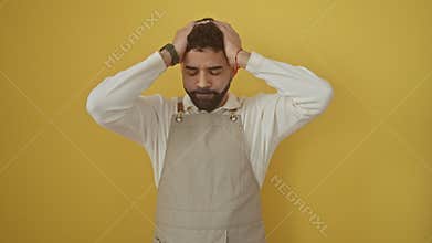 Young hispanic man in apron, suffering from stress-induced migraine, standing over yellow isolated background with aching hands on