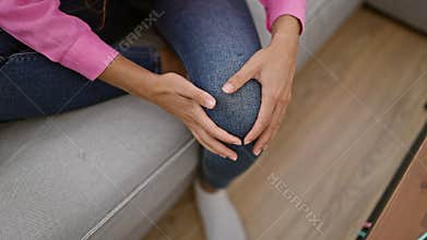 Hispanic woman\'s hands clutching aching knee, indoor suffering from a painful injury on living room sofa