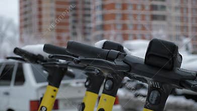 Close-up urban rental scooters stand in a row covered with snow on the road.