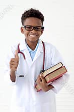 Portrait of happy smiling African boy in lab coat with stethoscope wearing glasses, holding pile of books on white wall room, cute