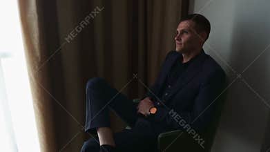 A man in a dark blue suit sits in an armchair in a window chair.