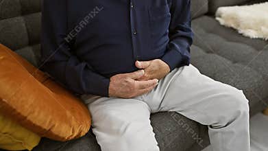 Senior man touching his aching stomach, suffering in pain at home, worriedly resting in the indoor sunlight
