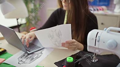 Focused woman sewing clothes in atelier with laptop, fabrics, mannequin, and sketch