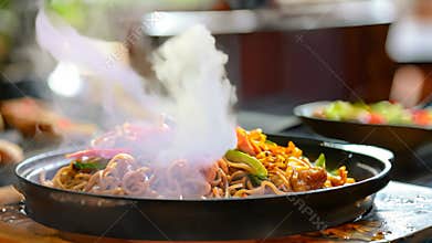 A wok filled with sizzling stir-fried noodles and colorful vegetables placed on a wooden table, A sizzling plate of stir-fried
