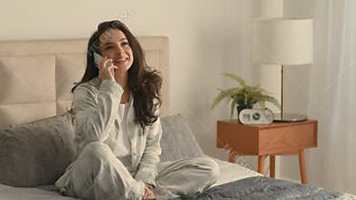Young Caucasian European woman sitting on bed home comfort weekend call picking up mobile phone smartphone telephone
