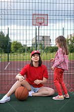 A child tries on his baseball cap to mother while sitting at the metal fence of basketball court