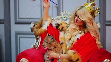 Blonde in red cloak with fur and queens crown sits