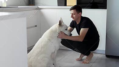 A young man is training a white Swiss shepherd dog in the kitchen. Pets concept. Leisure time with a pet