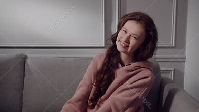 A happy European or American young lady sits on the sofa, squeezes shoulders, smiles and laughs. A girl teenager model