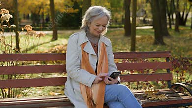 Happy grey-haired beautiful mature woman using smartphone online in autumn park Caucasian female using internet