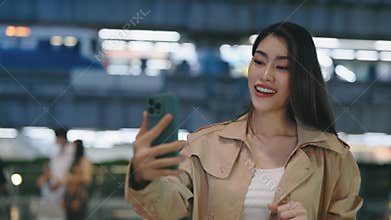 Charming Asian Girl Using Cell Phone for Video Call Standing in Night City Street with Lights on Background.