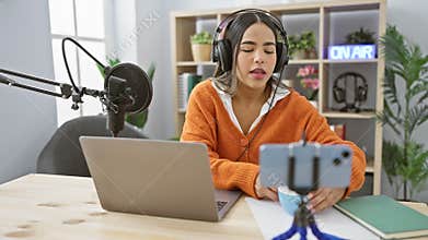 A young hispanic woman creates content in a studio, podcasting and streaming with a microphone, headphones, and laptop
