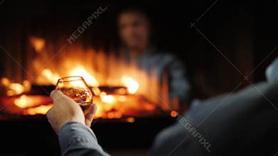 A man drinks brandy by the fireplace, his face is reflected in the glass of the furnace