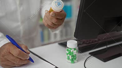 A senior male pharmacist writing a prescription in a pharmacy while holding two bottles of medication at a counter beside a