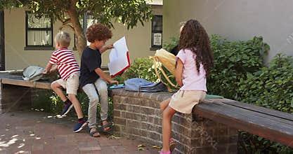 Three schoolchildren sitting on wall at school during break, sorting backpacks, opening red binder