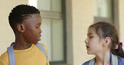 Diverse kids boy in yellow shirt glancing, speaking, girl in denim replying before entry backpacks