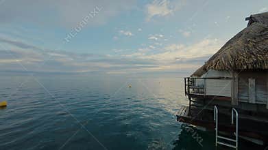 Tranquil ocean view at sunrise with a thatched-roof bungalow on stilts over calm blue water and sky