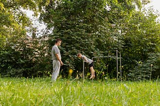 A father encourages his child as they play together on a playground. The child is attempting to perform a gymnastic move while