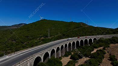 Drone view of traffic on arched Tarifa Bridge between green hills with wind turbines in distance