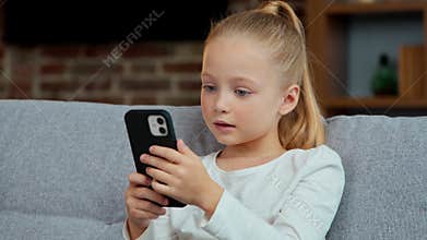 Caucasian little girl kid child daughter looking in mobile phone on sofa at home using smartphone play internet games