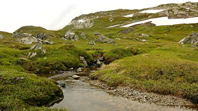 Small mountain stream in Alps, water is running over stones in fresh green meadow. High alps peaks in background.