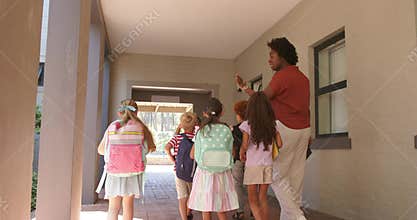 African American woman raising hand with tablet leading kids with backpacks down hall to courtyard