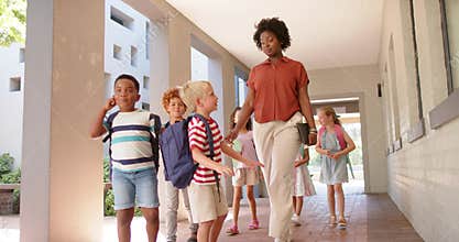 Female teacher with tablet leading children with backpacks hearing child gesturing guiding to class