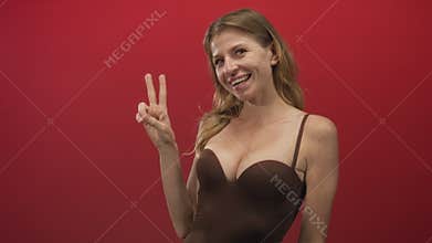 Woman with visible breasts smiling and flashing a two finger peace sign in a red studio setting; confidence peace