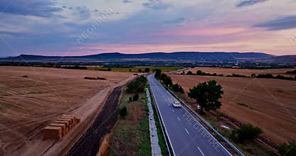 Fast Modern White Car Driving On a Rural Highway Through Agricultural Fields in the Countryside Under a Clear Blue Sky at Sunset.