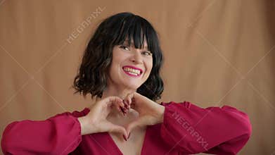 Bright smiling woman forms heart with fingers near chest. Gesture and warm smile express gratitude affection, tender emotion