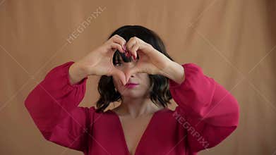 Playful woman forming heart shapes with hands near eyes and smiling in fashion red blouse. Gesture and wide happy smile convey