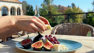 Hand reaches for cheese figs and grapes on a plate with wine bottle on a sunny outdoor table