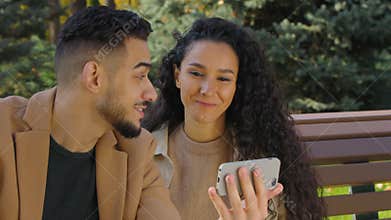 Happy hispanic couple talking video call with mobile phone in autumn city park sitting on bench family smiling Arabian