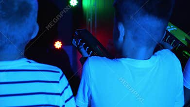 Teenage children shoot laser weapons at colored targets in the dark playroom.