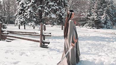 Charming brunette stands alone at the exit from her village, lowers her sword in snow and looks sad down, militant