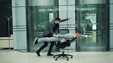 Funny young men are having fun with rolling chair, one is pushing armchair other is lying in it and moving forward in