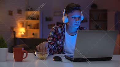 Overemotional teen playing computer game on laptop and eating snacks, addiction