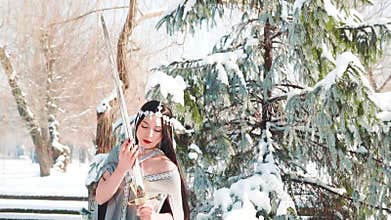 Pretty young lady in long gray cloak and dress in the snow and winter forest, the snow queen with silver sword in her