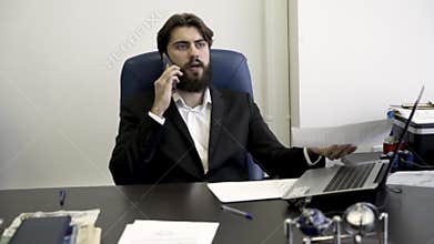 Concentrated, nervous, young, bearded businessman on the phone, sitting in blue leather armchair in the office. Angry