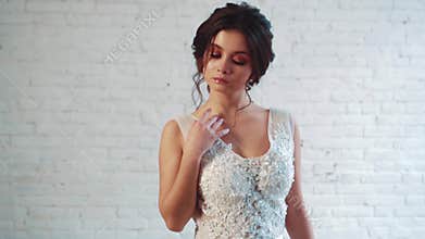 Sweet lady with dark hair in a gentle hairstyle is standing in the studio in a long elegant white dress, posing, raising