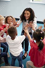 Elevated view of infant school children in a circle in the classroom giving high fives to their smiling female teacher, vertical,