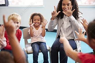 Front view of infant school children sitting on chairs in a circle in the classroom, holding up their hands and learning to count
