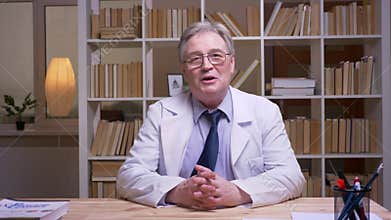 Interview of senior doctor in white coat talking into camera on book shelves background.